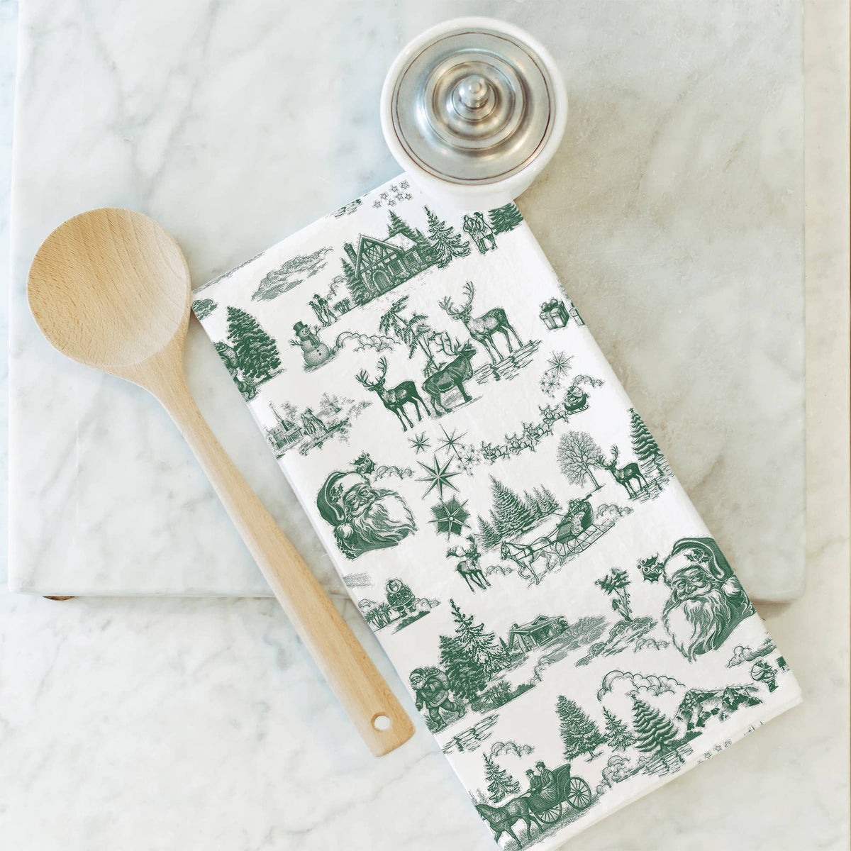 A white kitchen towel with a green Christmas-themed toile de Jouy pattern, styled on a white marble surface next to a wooden spoon and a small metal lidded container.