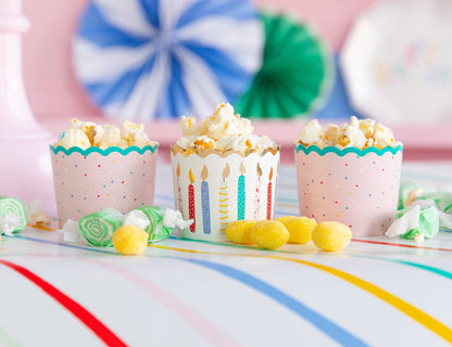 Three popcorn cups with colorful designs on a table with candy and party decorations.