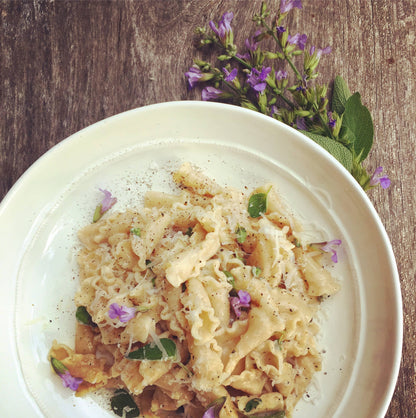 A close-up shot of a creamy pasta dish served on a white plate, garnished with herbs and small purple flowers, set on a rustic wooden table with a sprig of fresh herbs in the corner.