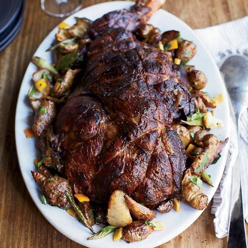 A large roasted lamb leg served on a white oval platter with roasted potatoes, green beans, and mustard accents, set on a wooden table with a napkin and cutlery nearby.
