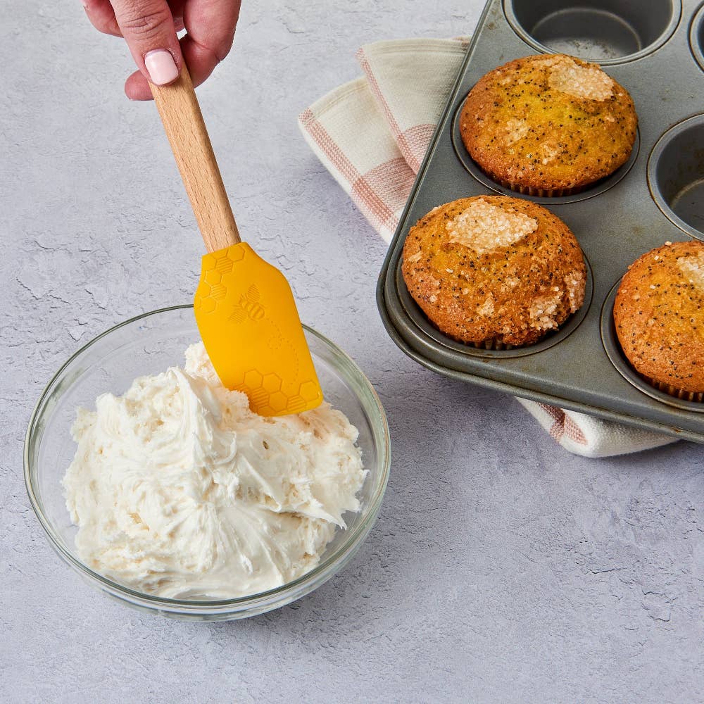 Bowl of cream cheese frosting with a spatula, muffin tin with muffins, and kitchen towel on a gray surface.