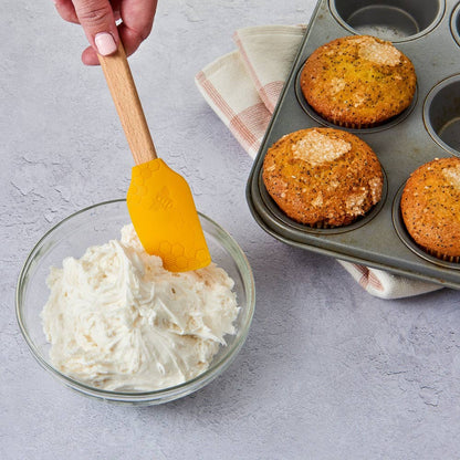 Bowl of cream cheese frosting with a spatula, muffin tin with muffins, and kitchen towel on a gray surface.