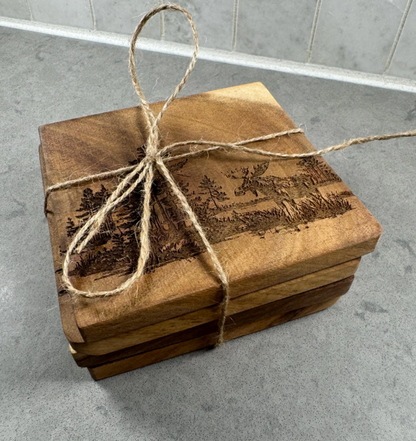 A stack of square wooden coasters with engraved landscape scenes, tied together with natural jute twine on a gray countertop.
