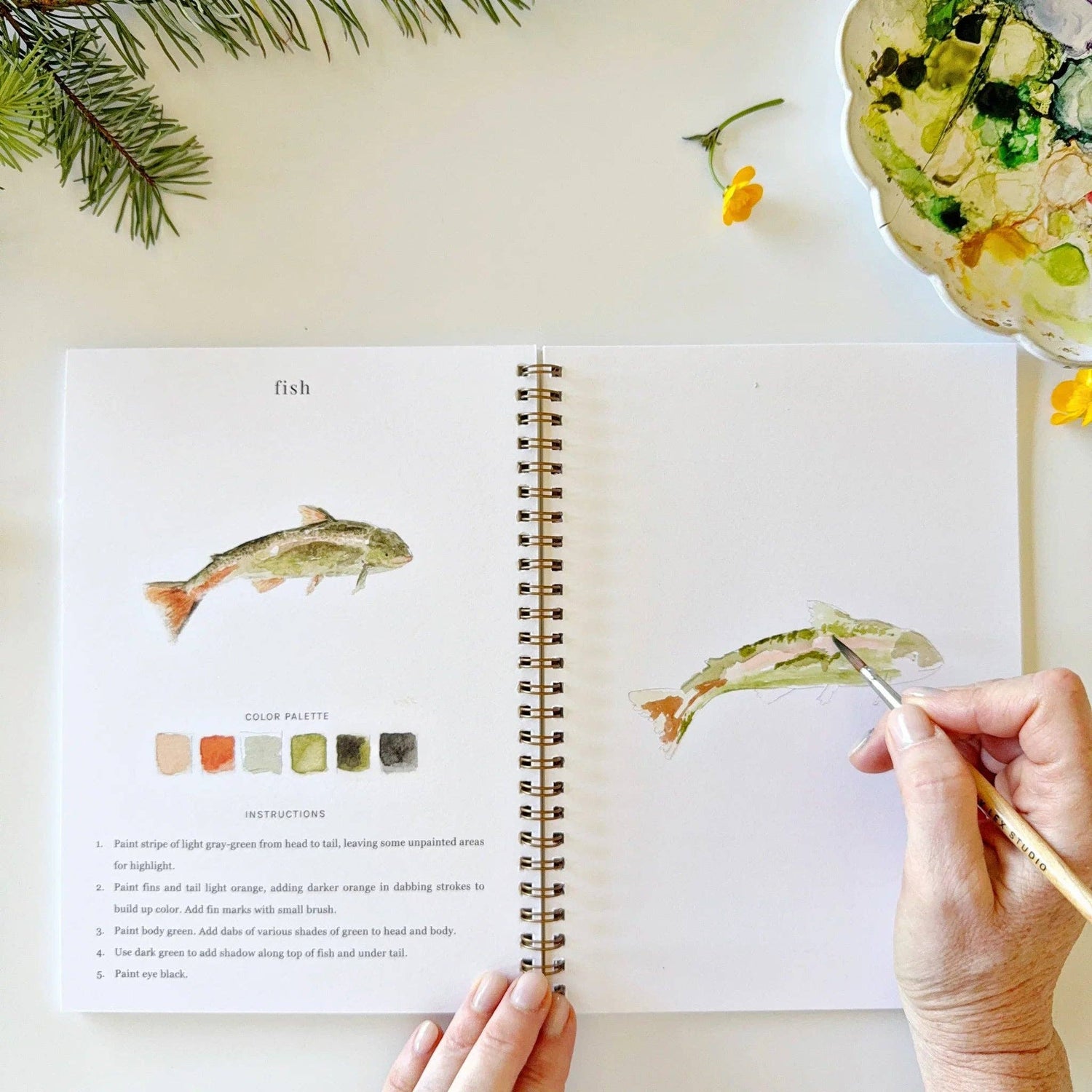 Open book with watercolor illustrations of fish, hand holding a paintbrush, and floral elements on a light background.