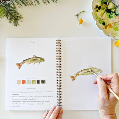 Open book with watercolor illustrations of fish, hand holding a paintbrush, and floral elements on a light background.