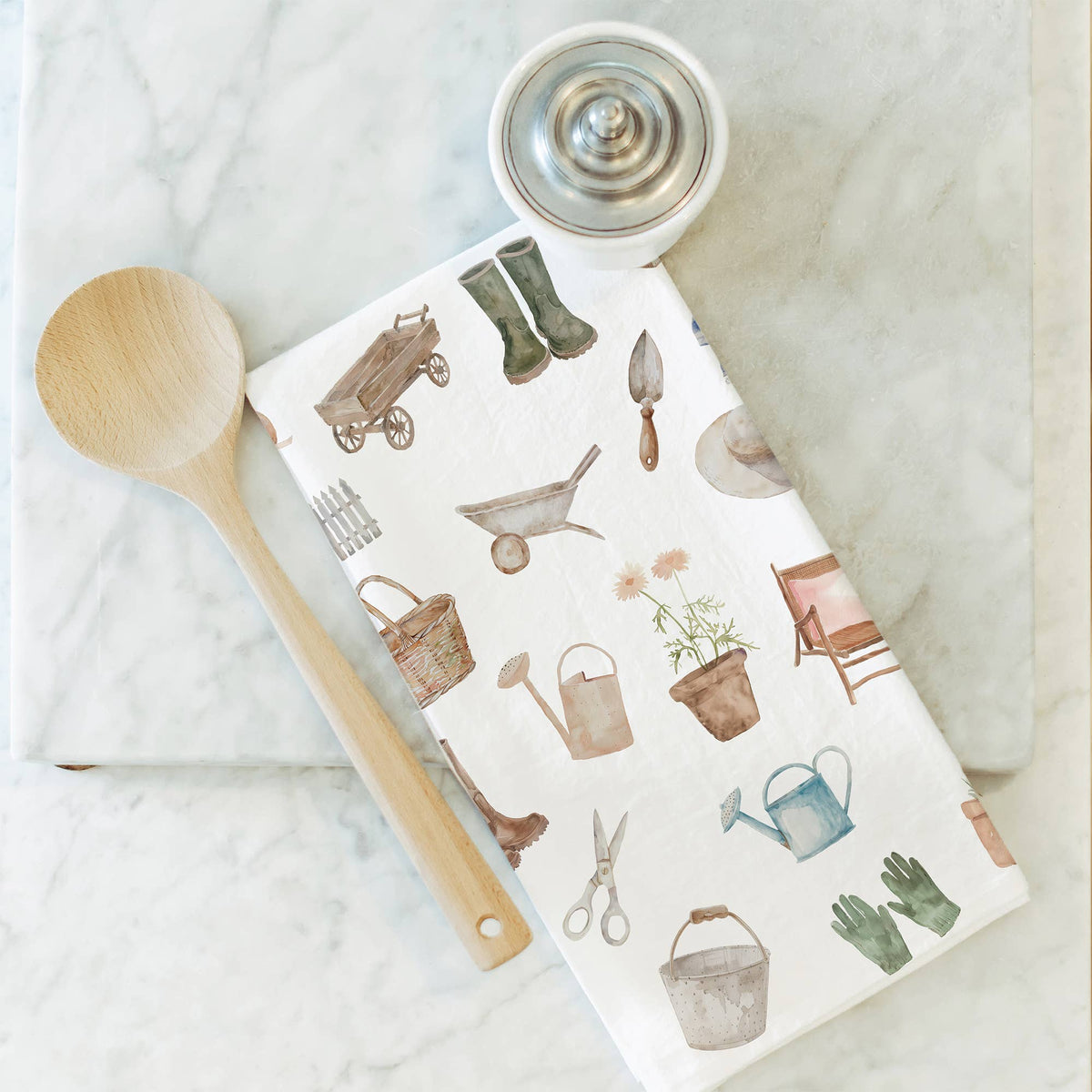Gardening-themed kitchen towel with tools and plants on a marble surface with a wooden spoon.