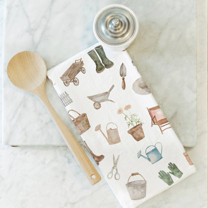 Gardening-themed kitchen towel with tools and plants on a marble surface with a wooden spoon.
