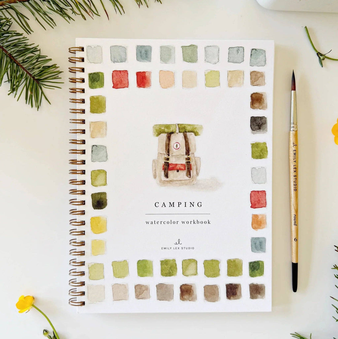 Camping watercolor workbook with a backpack illustration on a white surface with greenery.
