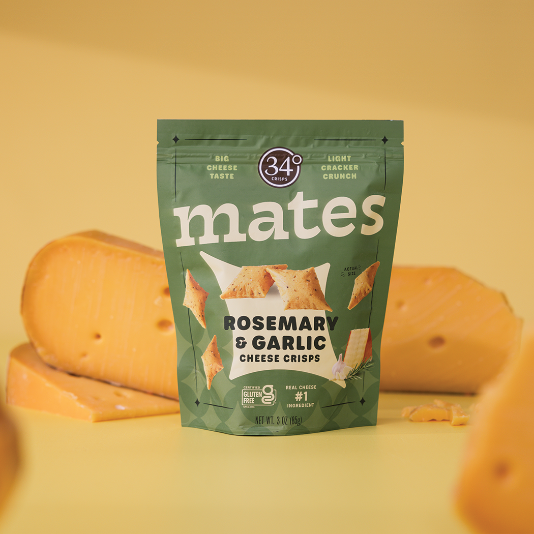 A package of 34° Cmates Rosemary &amp; Garlic Cheese Crisps stands centered against a warm yellow background with cheese wedges blurred in the foreground and behind it.