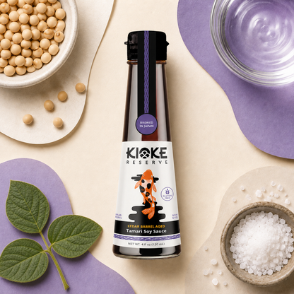 A bottle of Kioke Reserve Tamari Soy Sauce is centered in a styled flat-lay composition with bowls of soybeans, salt, water, and green leaves on a beige and purple background.