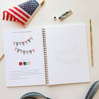 Open book with a stars and stripes banner design on a white surface with art supplies.