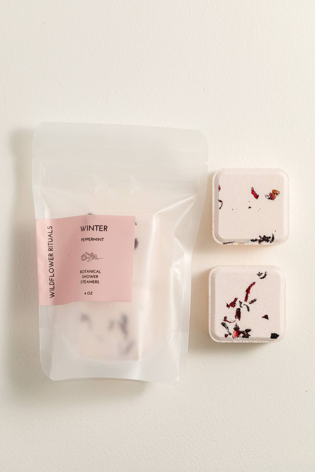 Two square shower steamers with floral designs on a white background, accompanied by a pink packaging labeled &