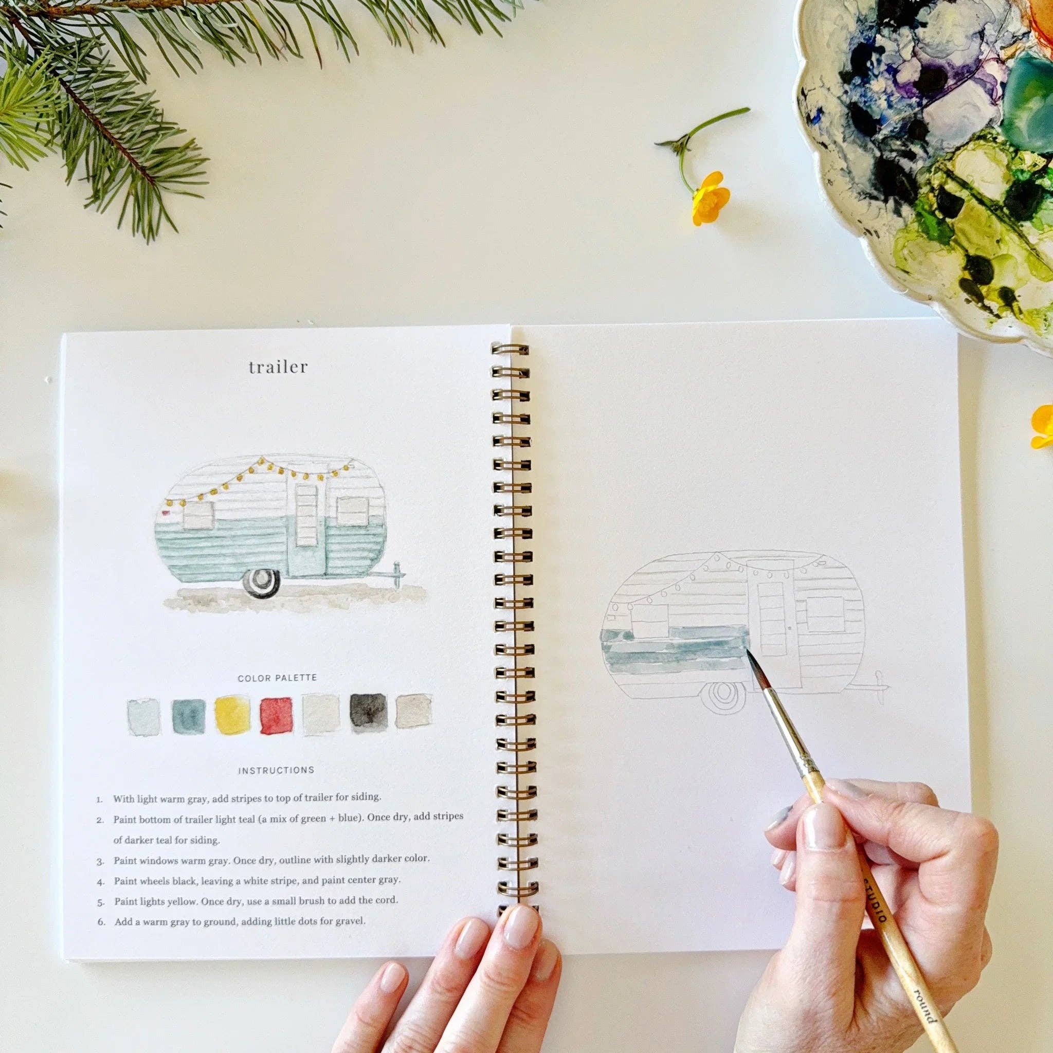 Open book with art project instructions, hand holding a paintbrush, and decorative elements on a light surface.