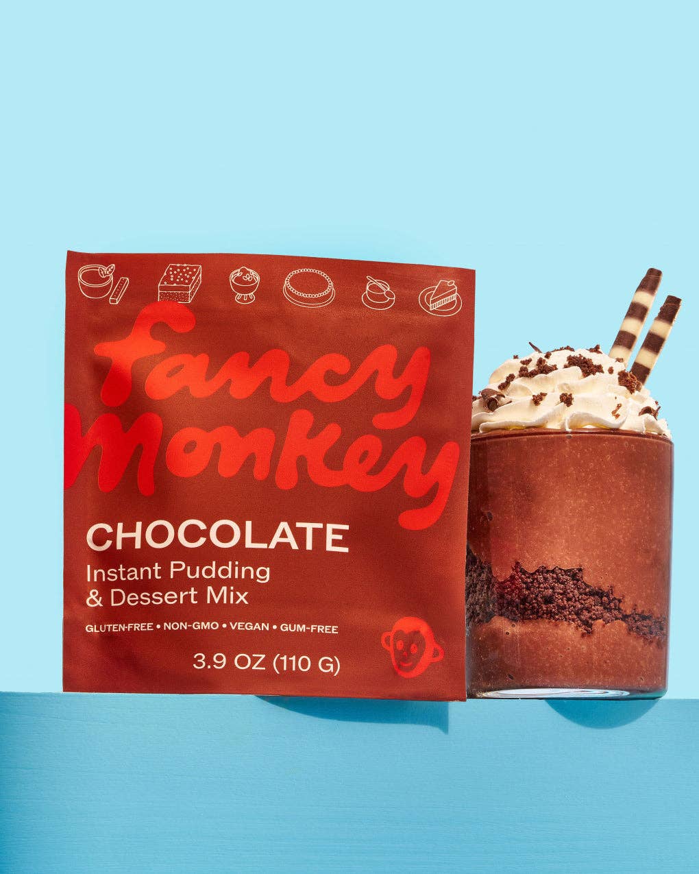 Fancy Monkey Chocolate Instant Pudding &amp; Dessert Mix packaging box next to a chocolate pudding drink topped with whipped cream and two striped straws on a light blue background.