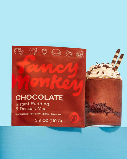 Fancy Monkey Chocolate Instant Pudding &amp; Dessert Mix packaging box next to a chocolate pudding drink topped with whipped cream and two striped straws on a light blue background.