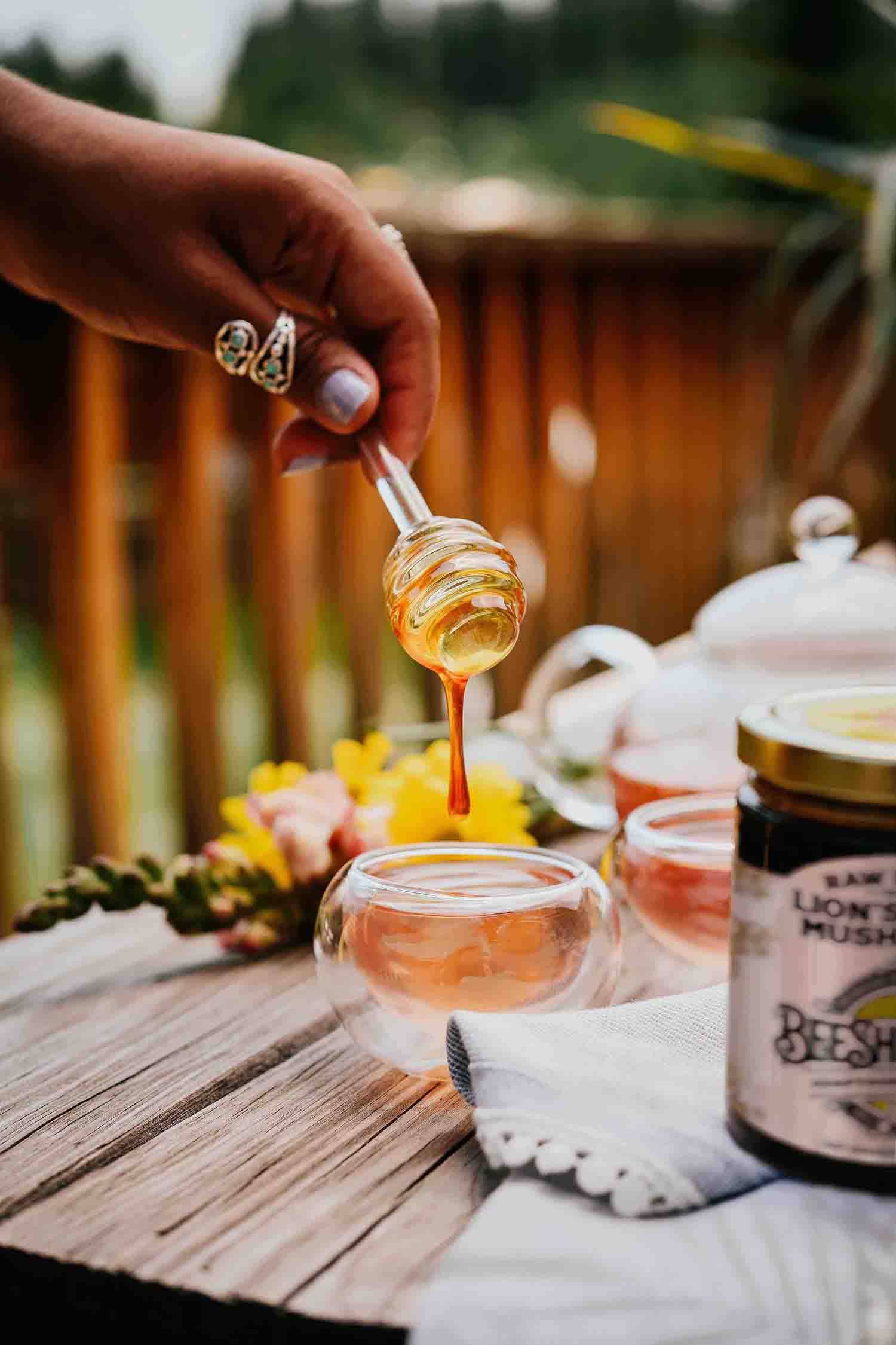 Person drizzling honey into a cup on a wooden table with tea and a jar of honey.