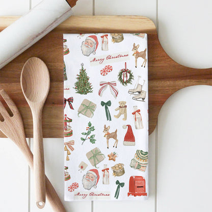 A white kitchen tea towel with a festive Christmas print, laid flat on a wooden cutting board with wooden spoons and a rolling pin nearby.