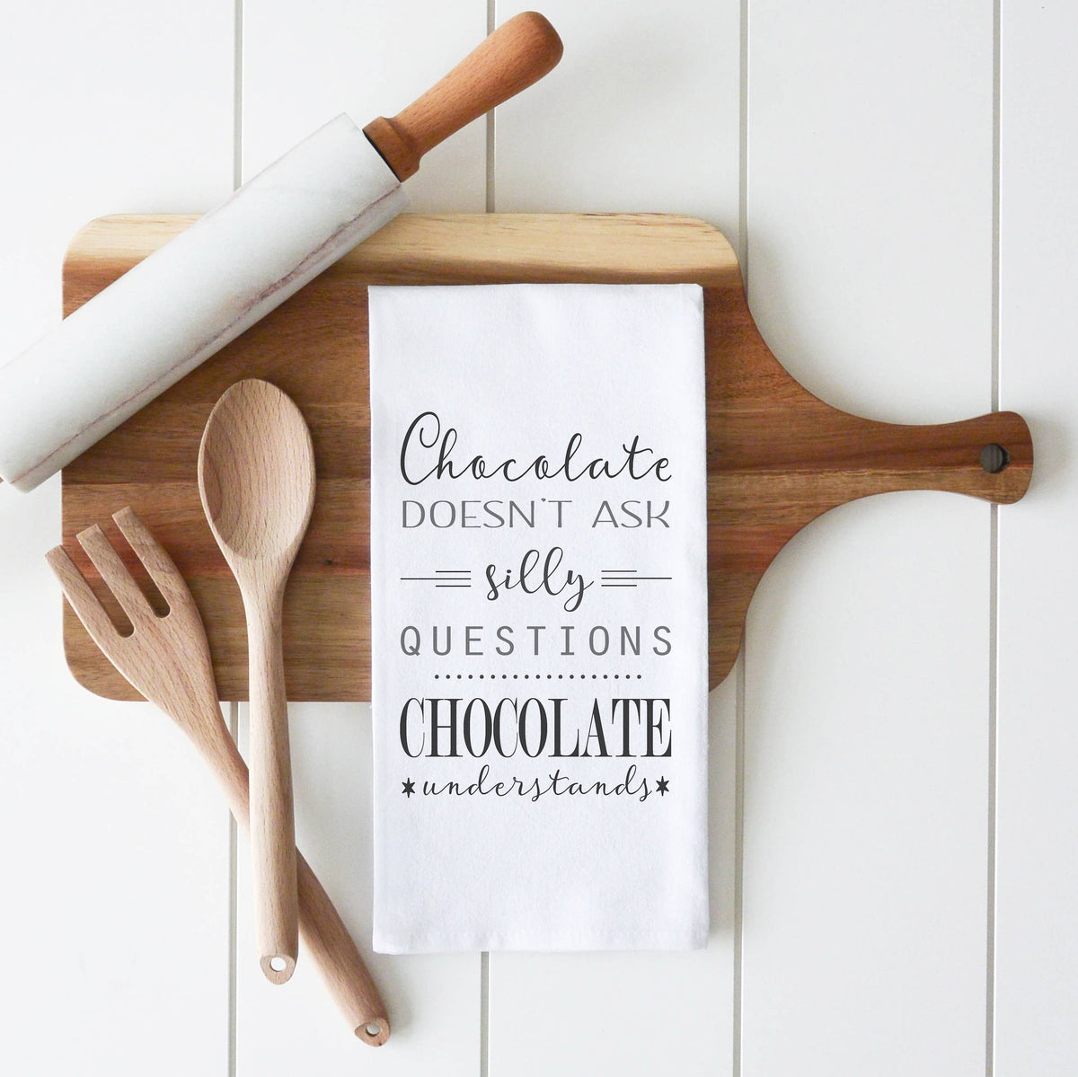 Wooden cutting board with kitchen utensils and a towel with a chocolate-themed quote on a white background
