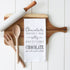 Wooden cutting board with kitchen utensils and a towel with a chocolate-themed quote on a white background