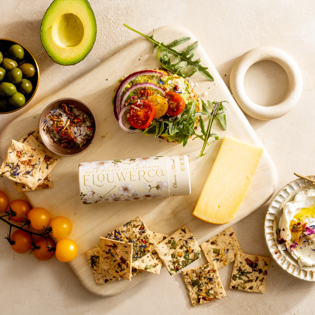 Platter with a sandwich, crackers, cheese, and a bottle of flower co product on a light background