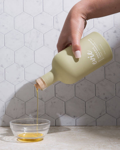 Hand pouring a liquid from a green bottle into a glass bowl against a tiled wall background