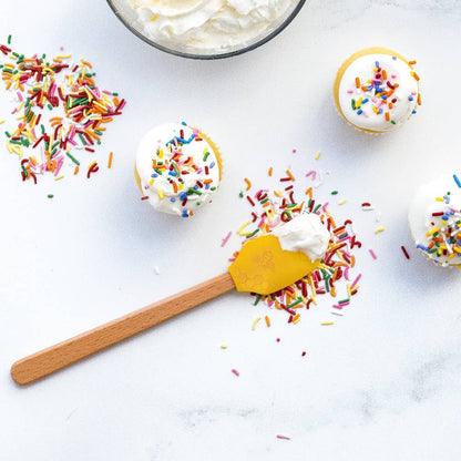 Cupcakes with white frosting and colorful sprinkles on a white surface with a yellow spatula.