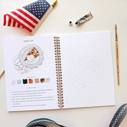 Open book with a drawing of apple pie on a white surface with art supplies.