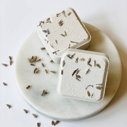 Two square shower steamers with dried herbs on a marble plate with scattered herbs on a light background