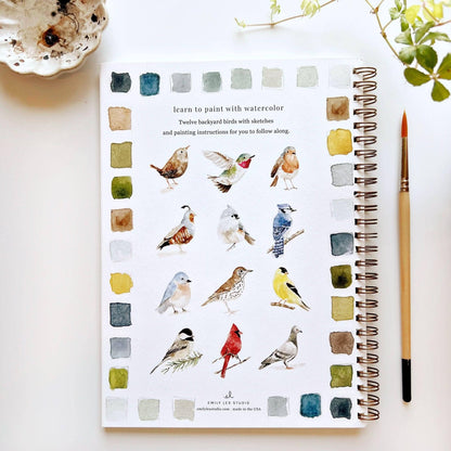 Notebook with watercolor illustrations of birds and a paintbrush on a white surface