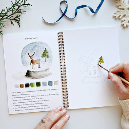 Person painting a snow globe illustration in a book with a paintbrush.