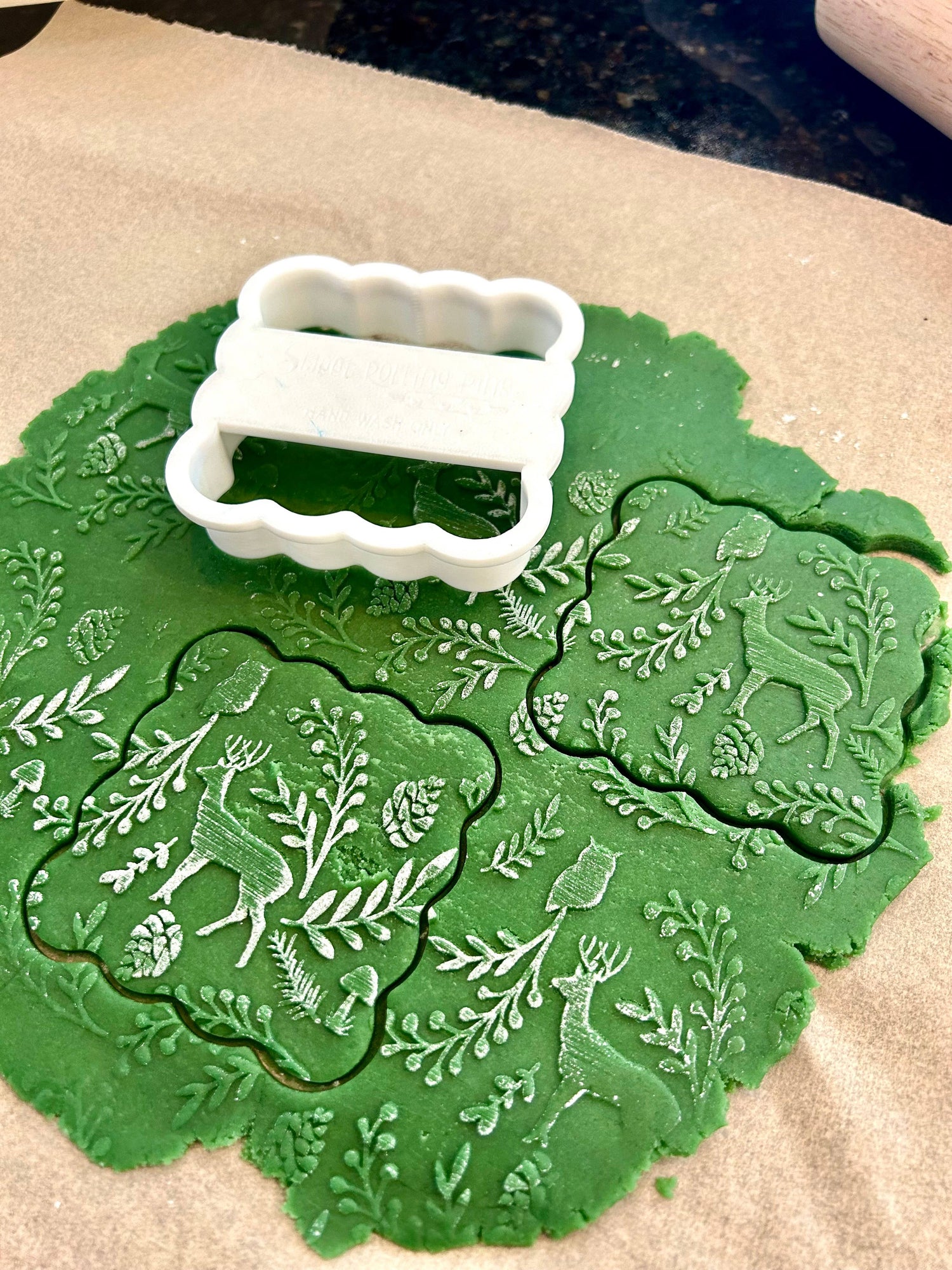 A white scalloped cookie cutter resting on top of green stamped dough on parchment paper. The green dough features embossed white designs of deer, pinecones, and leafy branches.