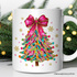 White mug with a colorful Christmas tree design featuring a pink bow, surrounded by festive greenery.
