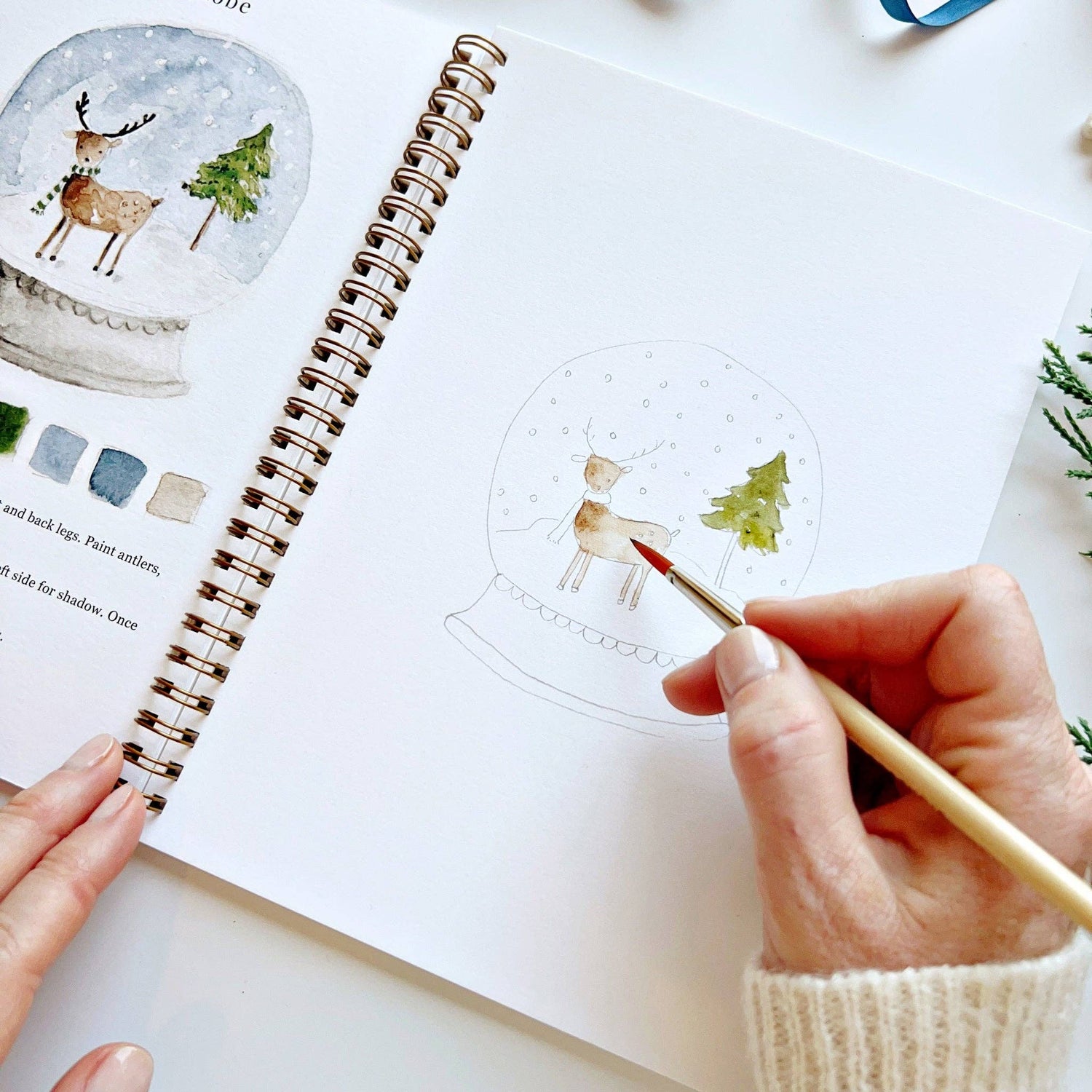Person coloring a reindeer in a snow globe illustration in a notebook.