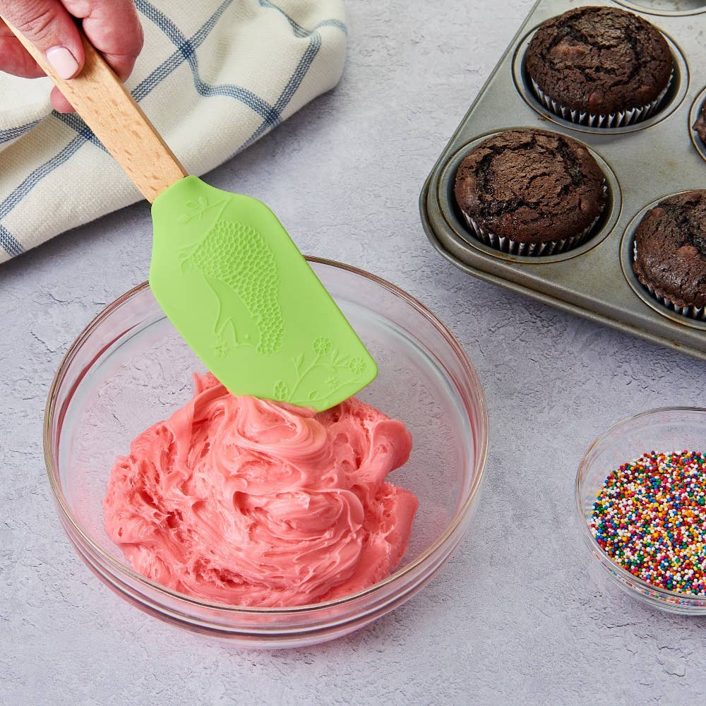 Pink frosting in a glass bowl with a green spatula, next to a muffin tin with cupcakes and a bowl of sprinkles.