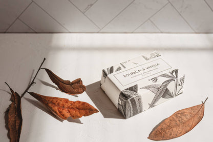 Bar of soap with packaging on a white surface with dried leaves and a branch.