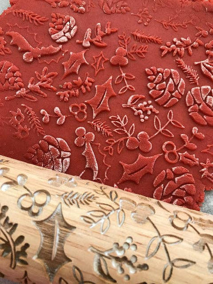 A close-up of a wooden rolling pin with engraved floral and botanical motifs resting over red stamped clay or fabric featuring matching red leaf and berry patterns.