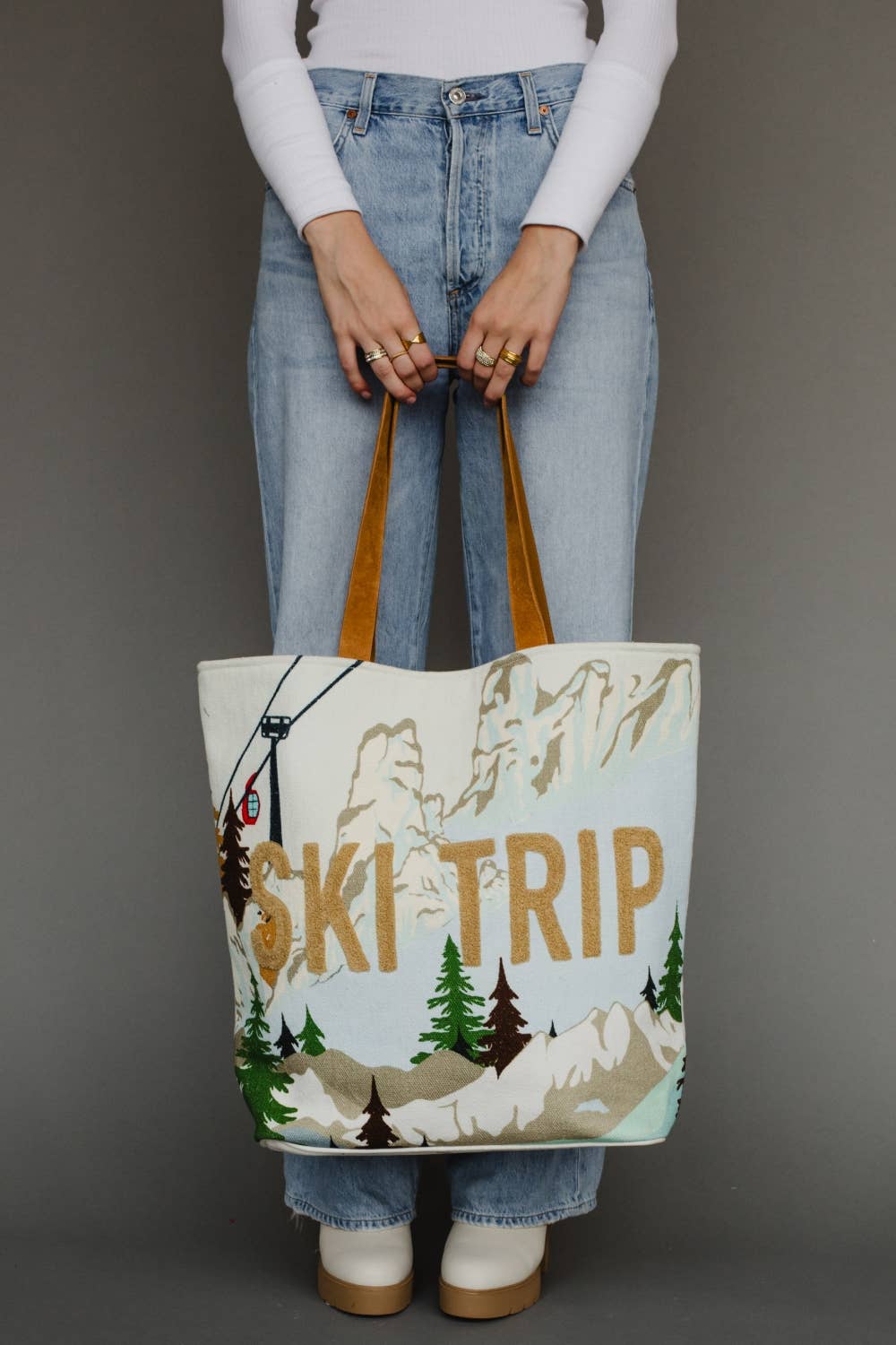 A person wearing light blue jeans and a white long-sleeve top holds a large canvas tote bag with brown handles. The bag has a winter mountain scene printed on it with the words &