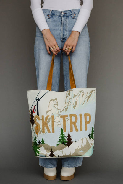 A person wearing light blue jeans and a white long-sleeve top holds a large canvas tote bag with brown handles. The bag has a winter mountain scene printed on it with the words &
