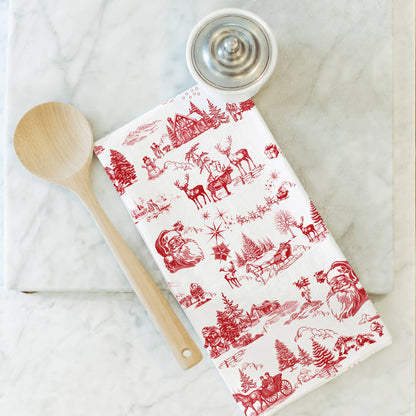 A white kitchen towel with a red Christmas-themed toile de Jouy pattern, styled on a white marble surface with a wooden spoon and a small metal container.