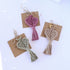 Three macrame earrings on brown paper tags with a plant in the background.
