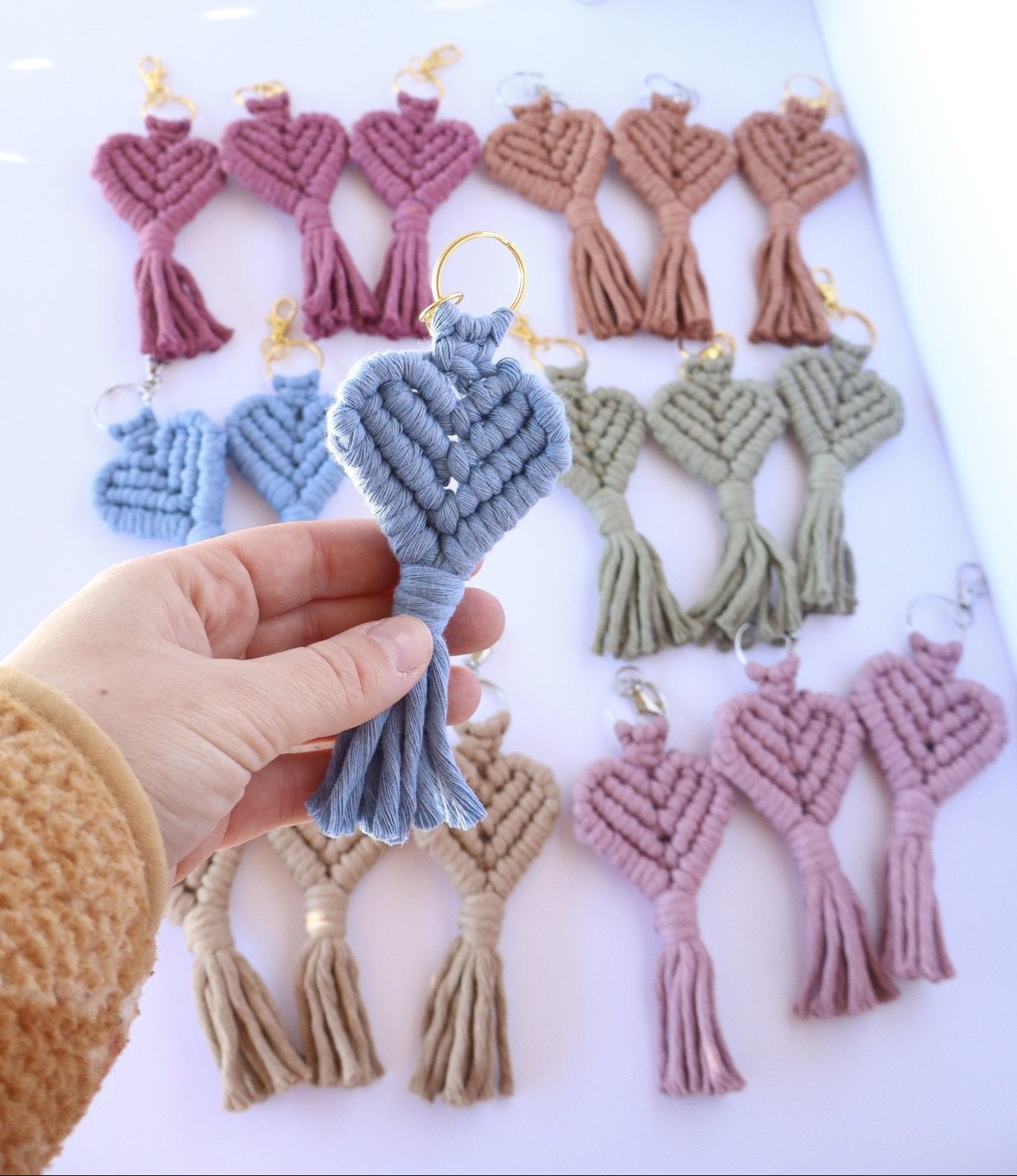Hand holding a blue macrame heart keychain with more keychains in the background on a white surface.